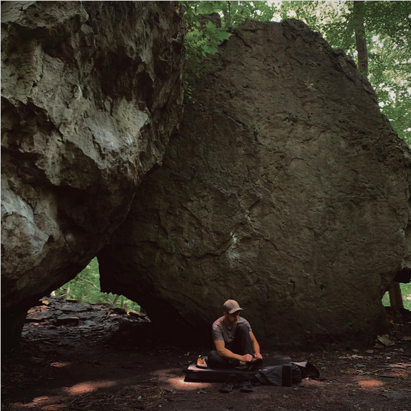 A picture of me putting on rock climbing shoes while sitting on a crash pad, a large boulder sits in the background with chalked up hand-holds